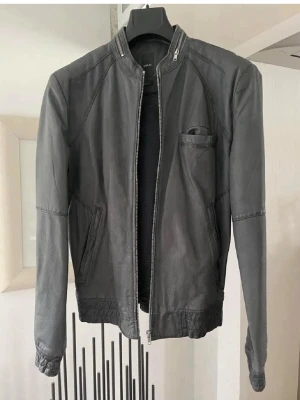 Grå  skinnjacka med dragkedja - Snygg grå skinnjacka med rak passform och dragkedja framtill. Jackan har två snedställda fickor och en liten bröstficka. Kragen är enkel utan slag och nederkanten har resårdetaljer. Perfekt för en cool och stilren look.