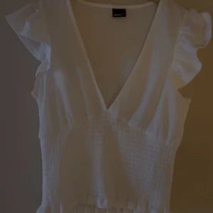 Vit omlottopp med volang från Gina Tricot - Söt vit omlottopp från Gina Tricot med volangdetaljer på ärmarna och smockad midja. Toppen har v-ringning och är tillverkad i ett lätt, luftigt material som passar perfekt till sommaren. Perfekt för dig som gillar feminina detaljer och en fräsch look.