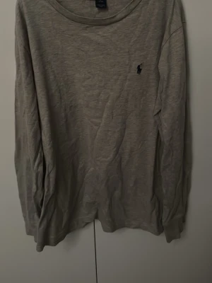 Grå långärmad T-shirt från Polo Ralph Lauren - Säljer en stilren grå långärmad T-shirt från Polo Ralph Lauren med den klassiska loggan broderad på bröstet. Tröjan har rund halsringning och är gjord i mjukt bomullsmaterial. Perfekt för en avslappnad och snygg look.