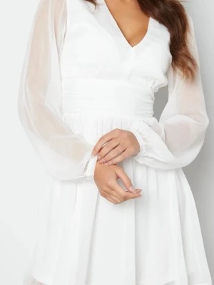 Vit kort omlottklänning med v-ringning - Söt vit kortklänning med v-ringning och transparenta långärmar. Klänningen har omlottdetalj i midjan och ett lätt, flödande tyg som ger en luftig känsla. Perfekt för studenten.