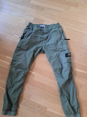 Olivgröna cargobyxor från Stone Island - Säljer ett par olivgröna cargobyxor från Stone Island med klassisk patch på benet. Byxorna har flera fickor, resår i midjan och benslut samt en avslappnad passform. Perfekta för en streetwear-look och riktigt snygga till sneakers.