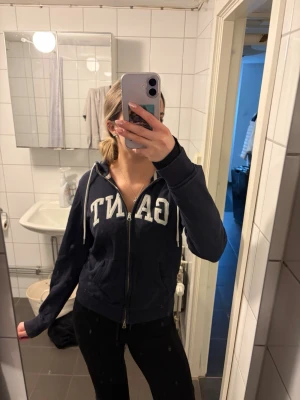 Gant zip hoodie – blå med vitt tryck - • Blå zip hoodie från GANT. • Vitt GANT-tryck över hela bröstet. • Vita dragsnören i huvan. • Dragkedja framtill. • Mjuk och bekväm hoodie. • Passar till vardag / casual.