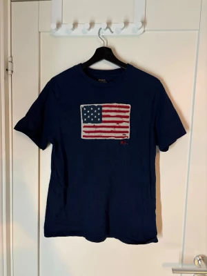 Mörkblå Polo Ralph Lauren t-shirt - Mörkblå t-shirt från Polo Ralph Lauren med ett stort tryck av den amerikanska flaggan på bröstet. Klassisk rund halsringning och korta ärmar. Perfekt för dig som gillar stilrena och ikoniska plagg. Pris kan diskuteras 