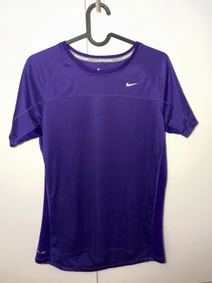 Lila Nike Dri-Fit löpartröja - Snygg lila löpartröja från Nike med Dri-Fit material som andas och transporterar bort fukt. Kortärmad modell med rund hals och diskret Nike-logga på bröstet. Perfekt för löpning eller träning.