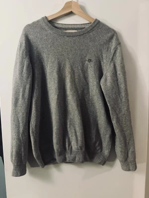 Grå stickad tröja från GANT - Klassisk grå stickad tröja från GANT i superfin lammull. Tröjan har rund halsringning, ribbade muddar och en diskret broderad logga på bröstet. Perfekt för lager-på-lager och enkel att matcha med jeans eller kjol.