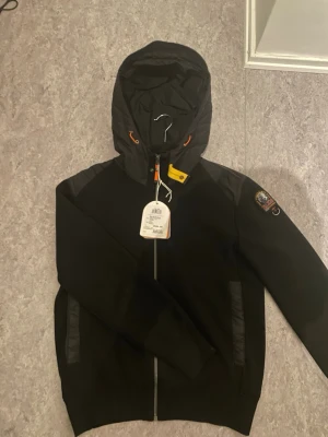 Parajumper cardigan - Svart cardigan från Parajumpers med huva, dragkedja framtill och detaljer i orange och gult. Jackan har en patch på ärmen och förstärkta partier på axlarna. Perfekt för dig som vill ha en stilren och funktionell jacka.