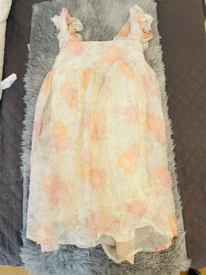 Blommig sommarklänning med volang - Söt kortklänning med volangdetaljer på axelbanden och blommigt mönster i pastellfärger. Klänningen har en rosett i ryggen och är tillverkad i ett lätt, luftigt material som passar perfekt för varma dagar.