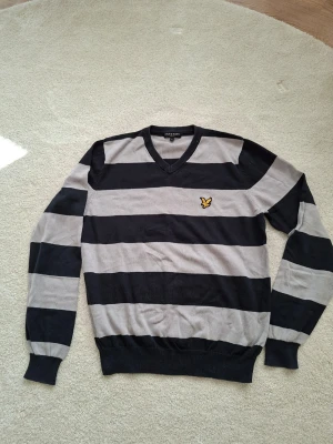 Randig stickad tröja från Lyle & Scott - Snygg stickad tröja med breda svarta och grå ränder från Lyle & Scott. V-ringad modell med lång ärm och klassisk gul logga på bröstet. Perfekt för en stilren och avslappnad look.