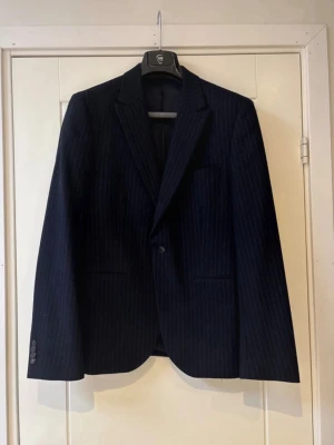 Mörkblå randig kavaj från Bläck - Snygg mörkblå kavaj från Bläck med diskreta vita ränder. Klassisk modell med två framfickor, bröstficka och knappar vid ärmslut. Regular fit och helfodrad insida för en clean look. Perfekt till skjorta och jeans eller kostymbyxor.