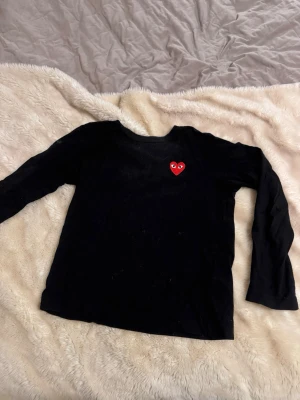 Svart långärmad tröja Comme des Garçons- play - Svart långärmad tröja från Comme des Garçons Play med det ikoniska röda hjärtat broderat på bröstet. Klassisk rund halsringning och mjukt bomullsmaterial. Perfekt för en clean och stilren look. Strl M men sitter som en s