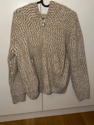 Beige stickad hoodie med dragkedja - Mysig stickad hoodie i beige och gråmelerat mönster. Tröjan har dragkedja framtill, huva och ribbstickade muddar vid ärmslut och nederkant. Perfekt för lager-på-lager och chill dagar.