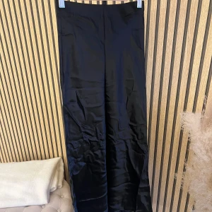 Svart satin kjol från h&m - Säljer en svart långkjol i glansigt satinmaterial. Kjolen har en rak och enkel siluett som ger en clean look. Perfekt för dig som gillar minimalistisk stil och vill ha något som känns både trendigt och klassiskt.