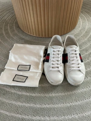 Gucci Ace  - Säljer ett par klassiska Gucci Ace sneakers i vitt läder med de ikoniska röda och mörkblå ränderna på sidorna. Skorna har svarta detaljer vid hälen och vita snören. Kommer med original dustbags. Perfekta för dig som gillar lyxiga och stilrena sneakers.