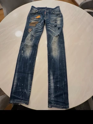 Blåa dsqduared - Säljer ett par blå jeans med coola patchar och slitningar, samt färgstänk för en trendig look. Jeansen har raka ben och flera detaljerade broderade märken på ena låret. Perfekta för dig som gillar streetstyle och vill sticka ut.
