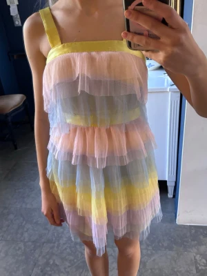 Pastellfärgad volangklänning - Har hål i sidan från förra ägare i syfte att trä band igenom och knyta för att kunna ha klänningen lösare/tajtare. Söt kortklänning i pastellfärger med flera lager volanger i rosa, blått, gult och lila.