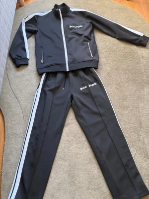 Palm Angels svart träningsset - Svart träningsset från Palm Angels med vita detaljer och logga på både jacka och byxa. Setet har dragkedja framtill, fickor med dragkedja och vita ränder längs ärmar och ben. Perfekt för sport eller chill. Materialet är troligtvis polyester.