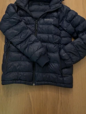 Mörkblå dunjacka från Peak Performance - Snygg mörkblå dunjacka från Peak Performance med huva och dragkedja framtill. Jackan har quiltad design, två sidofickor med dragkedja och muddar vid ärmsluten. Perfekt för kalla vinterdagar och riktigt skön att bära . Men på bild 6 och 7 så ser man att det finns två små hål vid vänstra armen men det syns inte när man har på jackan