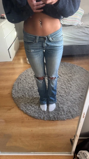 Ljusblå slitna bootcut jeans Levi's - Säljer ett par ljusblå bootcut jeans från Levi's med slitningar och hål vid knäna. Jeansen har klassiska bakfickor med gul kontrastsöm och Levi's röda etikett. Perfekta för en avslappnad och trendig look.