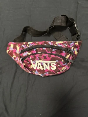 Färgglad magväska från Vans - Säljer en cool magväska från Vans med färgstarkt kamouflagemönster i rosa, lila, gul och brunt. Väskan har två dragkedjefack på framsidan och justerbart svart band med spänne. Perfekt för att bära småsaker och hålla händerna fria.