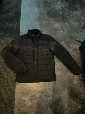 Marinblå vårjacka - Tunnare dunjacka med tryckknappar framtill. Jackan har två bröstfickor med lock och två sidofickor. Insidan har två innerfickor. Jackan har en hög krage som är perfekt för blåsiga vårdagar. Med en tunn och lätt dunstoppning passar den bra för den kommande våren! Hör av er vid några funderingar!!!