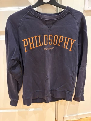 Mörkblå sweatshirt från Gant - Mörkblå sweatshirt från Gant med orange PHILOSOPHY-tryck på bröstet. Klassisk rund halsringning, ribbade muddar och avslappnad passform. Perfekt för en chill och stilren look.