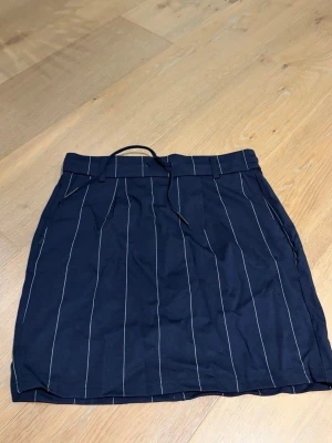 Mörkblå randig kjol från ONLY - Snygg mörkblå kortkjol från ONLY med vita vertikala ränder och knytband i midjan. Kjolen har en klassisk passform och är perfekt för dig som gillar en stilren look. Tillverkad i ett mjukt och bekvämt material.