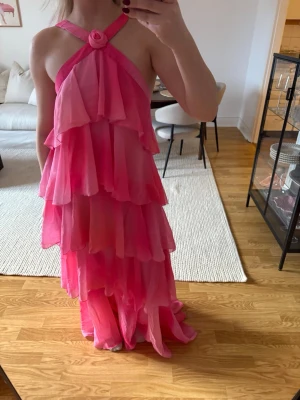 Rosa volangklänning med öppen rygg från Vero Moda - Säljer en långklänning i flera nyanser av rosa med volanger i lager på lager. Klänningen har halterneck med knut framtill och öppen rygg. Perfekt för dig som vill sticka ut med en färgstark och flowy look.