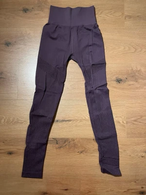 Mörklila seamless tights - Säljer ett par mörklila seamless tights med hög midja och ventilerande meshdetaljer längs benen. Perfekta för träning eller gymmet, med stretchigt material som ger bra rörelsefrihet. Snygg design med diskreta mönster på benen.