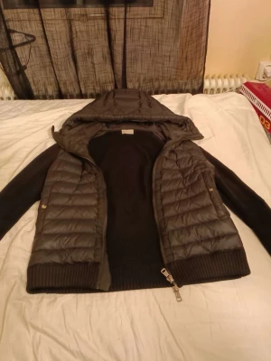 Svart hybrid-cardigan med huva - Snygg svart cardiganjacka med stickade ärmar och rygg samt vadderad framsida. Jackan har en praktisk huva och dragkedja framtill. Perfekt för lager-på-lager och streetwear-stil. Materialmix av stickat tyg och quiltad polyester. Kvitto finns, priset är inte hugget i sten 