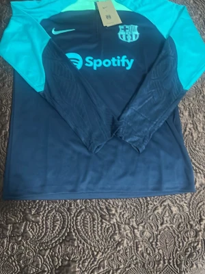 Barcelona träningströja Nike blå/turkos - Snygg långärmad träningströja från Nike för FC Barcelona i mörkblått med turkosa ärmar och detaljer. Tröjan har klubbmärke, Nike-logga och Spotify som sponsortryck på bröstet. Perfekt för fotbollsträning eller sportig stil. Om ni vill ha mer bilder fråga 