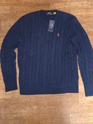 Mörkblå kabelstickad tröja Polo Ralph Lauren - Kabelstickad mörkblå tröja från Polo Ralph Lauren med rund halsringning och ribbade muddar. Tröjan har det klassiska röda Polo-logot broderat på bröstet och är tillverkad i ett mjukt material. Perfekt för en stilren och avslappnad look.
