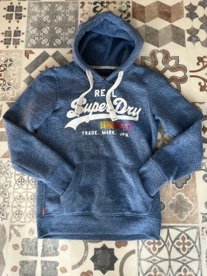 Blå Superdry hoodie med tryck - Säljer en blå hoodie från Superdry med vit logga och färgglatt japanskt tryck på bröstet. Hoodien har huva med vita snören och en stor magficka. Tillverkad i mjukt bomullsmaterial och har ribbade muddar vid ärmar och nederkant. Perfekt för en avslappnad streetstyle.