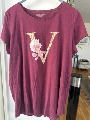 Vinröd sovtröja Victoria's Secret - Mysig vinröd sovtröja från Victoria's Secret med ett stort V i leopardmönster och en rosa blomma på bröstet. T-shirten har rund halsringning och korta ärmar, perfekt för chillkvällar hemma. Använd vid enstaka tillfällen och är i väldigt bra skick 