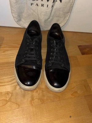 Svarta sneakers från Lanvin - Säljer ett par svarta sneakers från Lanvin med snörning och blank svart tå. Skorna har en vit sula och är tillverkade i mocka och skinn, vilket ger en lyxig känsla. Perfekta för dig som vill ha stilrena och trendiga sneakers.