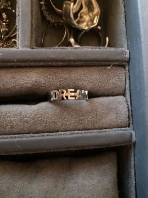 Snygg silverring med DREAM-text - Cool ring i silverfärgad metall med utskuren texten 'DREAM' runt hela ringen. Perfekt för dig som gillar statement-smycken och vill ha något unikt på fingret. Enkel men ändå iögonfallande design som passar till många olika stilar. Den är av rostfrittstål.