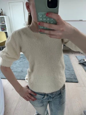 Krämvit fluffig stickad tröja Gina Tricot - Mysig krämvit stickad tröja från Gina Tricot med fluffig textur. Tröjan har rund halsringning, kortare ärmar och ribbade muddar vid ärmslut och nederkant. Perfekt för dig som gillar en enkel men trendig look.