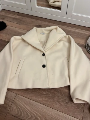 Krämvit kort kappa från Noisy May - Stilren krämvit kappa från Noisy May med kort, boxig passform och bred krage. Kappan har två svarta knappar framtill och snedställda fickor. Tillverkad i mjukt, ull-liknande material som ger en trendig och clean look.