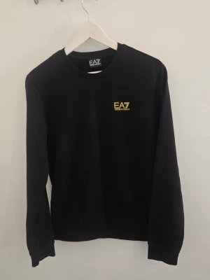 Svart sweatshirt från EA7 Emporio Armani - Svart sweatshirt med rund hals från EA7 Emporio Armani. Tröjan har gul logga på bröstet och gula ränder i nacken på baksidan. Långärmad modell i mjukt material, perfekt för en clean och stilren look. Prövad nångång