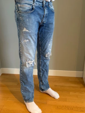 Replay Grover slitna - Säljer ett par blå jeans från Replay med slitningar och hål på knäna. Jeansen har klassisk femficksmodell, normal midja och raka ben. Materialet är denim i bomull och tvätten är ljusblå med tydliga slitningar för en trendig look. Är 185 och sitter bra