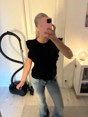 Svart blus med volangärmar - Säljer en svart blus med volangdetaljer på ärmarna. Blusen är ärmlös och har en rund halsringning. Passar perfekt till både jeans och kjol för en trendig look.