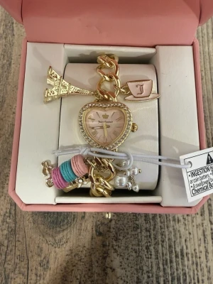 Guldig Juicy Couture charm-klocka - Unik guldig klocka från Juicy Couture med hjärtformat ur och gnistrande stenar runt boetten. Armbandet har chunky länkar och flera söta berlocker, bland annat Eiffeltornet, pärlor och färgglada detaljer i rosa, blått och beige. Perfekt accessoar för dig som gillar statement-smycken.