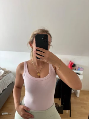Ljusrosa ribbad body - Säljer en ljusrosa bodysuit med breda axelband och rundad urringning. Materialet är ribbat och stretchigt, vilket ger en tight och bekväm passform. Perfekt att styla med jeans eller kjol för en enkel och snygg look.