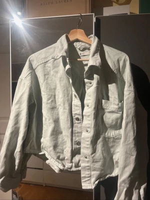 Ljus jeansjacka med cropped passform - Säljer en ljus jeansjacka med avslappnad, boxig och croppad passform. Jackan har klassisk krage, bröstficka och silverfärgade knappar framtill. Perfekt att slänga över en hoodie eller t-shirt för en chill look.