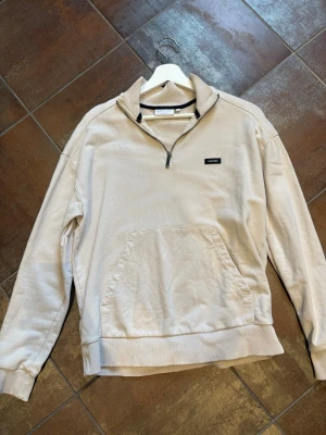 Calvin Klein halvzip - Snygg beige sweatshirt från Calvin Klein med halv dragkedja och hög krage. Tröjan har en stor magficka och ribbade muddar vid ärmslut och nederkant. Perfekt för en avslappnad och stilren look.