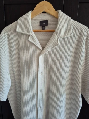 Hm vit ribbad skjorta  - Säljer en vit skjorta från H&M i loose fit med kort ärm och ribbad struktur. Skjortan är tillverkad i bomull och har klassisk krage samt knäppning framtill. Perfekt för en avslappnad och stilren look.