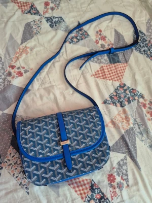 Goyard axelväska  - Snygg blå axelväska från Goyard med klassiskt mönster i vitt och grått. Väskan har justerbar axelrem och silverfärgad detalj framtill. Tillverkad i canvas med läderdetaljer. Perfekt för dig som vill ha en unik och trendig accessoar.