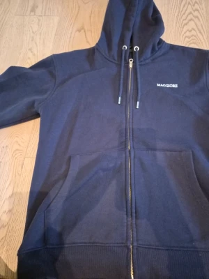 Maggiore Hoodie - Mörkblå zip-hoodie från Maggiore med en avslappnad och enkel design som passar perfekt för både vardag och fritid. Tröjan har huva med dragsko, fickor framtill och en normal passform. Mjuk fleece finns på insidan. Märkets logga är broderad i vitt och placerad på bröstet. Denna hoodie är lika snygg öppen som stängd.  Storlek M Ungdom