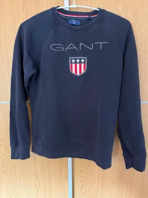 Mörkblå sweatshirt från GANT - Mörkblå sweatshirt från GANT med broderad logga och sköld med stjärnor och ränder på bröstet. Tröjan har rund halsringning och ribbade muddar vid ärmslut och nederkant. Perfekt för en avslappnad och stilren look.