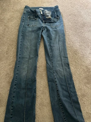 Blå bootcut jeans med söm - Säljer ett par blå bootcut jeans från Nelly med normal midja och dekorativa sömmar framtill. Jeansen har dubbla knappar, dragkedja och två fickor fram. Jeansen är i stretchigt material. Klassisk denimlook som passar perfekt till sneakers eller boots.