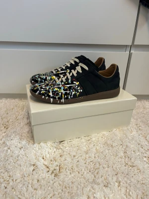 Svarta Maison margiela paint splatter gats - Unika svarta sneakers med färgstänk i vitt, gult och rött över tåpartiet. Skorna har vita/beiga snören, bruna sulor och insida i beige. Materialet ser ut att vara mocka eller nubuck. Perfekta för dig som vill sticka ut med din stil. Priset är inte fast skriv vid funderingar!